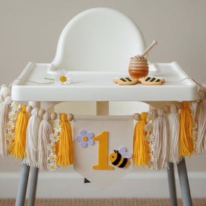 May include: A white high chair decorated with a banner and tassels. The banner has a large yellow number 1, a felt bee, and a purple flower. The tassels are yellow, cream, and beige. A small honey pot, cookies, and a daisy are on the tray.