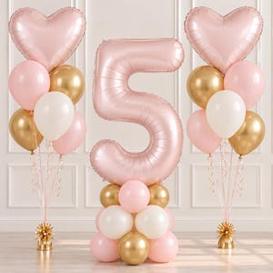 May include: A balloon arrangement featuring a large number five balloon in matte pink, flanked by two balloon bouquets. Each bouquet includes a heart-shaped pink balloon, round pink, white, and metallic gold balloons, and a gold base weight.