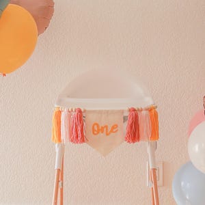 K&ouml;nnte beinhalten: Ein Hochstuhl, verziert mit einem Banner mit der Aufschrift "One". Das Banner ist mit orangefarbenen und rosa Quasten und Holzperlen verziert. Im Hintergrund sind pastellfarbene Luftballons zu sehen. Der Stuhl steht vor einer hellrosa Wand.