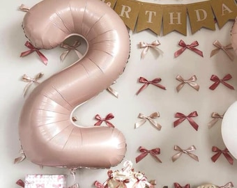 May include: A large, pink number 2 balloon is the focal point of this birthday celebration. The balloon is surrounded by decorative bows in shades of pink and cream. A small cake with pink bows and a banner that says "HAPPY BIRTHDAY" complete the festive scene.