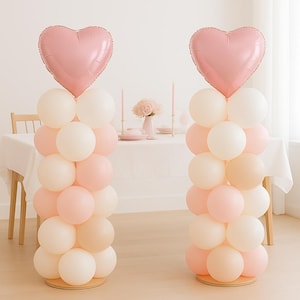 Puede incluir: Dos columnas de globos con un globo en forma de corazón en la parte superior. Las columnas están hechas de globos blancos, rosas y melocotón. Los globos de corazón son de color rosa claro. Las columnas están sobre una base de madera, con una mesa en el fondo.