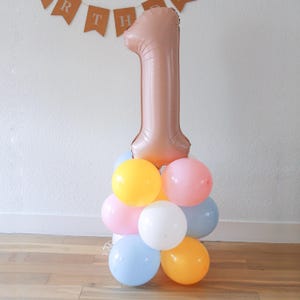 Peut inclure: Un grand ballon en forme de numéro un en or rose avec un groupe de ballons de couleur pastel en dessous. Les ballons sont roses, jaunes, bleus et blancs.