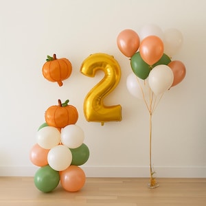 May include: A festive arrangement of balloons and decorations. Includes a large gold number 2 balloon, orange pumpkin-shaped balloons, and a cluster of balloons in white, green, and peach colors. Perfect for a second birthday celebration.