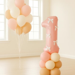 May include: A festive arrangement of balloons for a first birthday celebration. The display includes a large pink number one balloon adorned with white bows, and a cluster of pastel-colored balloons in pink, cream, and peach.