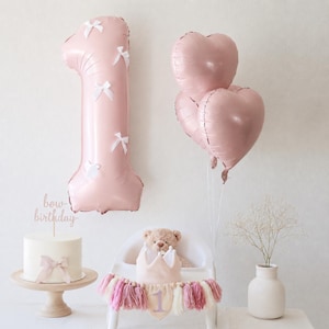 May include: A light pink number one balloon decorated with small white bows, three heart-shaped pink balloons, a white cake with a bow and a cake topper reading bow birthday, a teddy bear, a crown, and a tassel banner on a high chair.