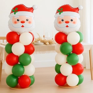 May include: Two Christmas balloon decorations. Each features a Santa Claus head balloon atop a column of red, green, and white balloons. The balloons are arranged on a round wooden base, creating festive holiday decor.
