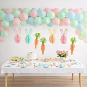 May include: A pastel-themed Easter table setting. A white tablecloth covers a wooden table, set with plates, cutlery, and treats. Above, a balloon arch in pastel colours and hanging paper carrots and bunny decorations complete the festive scene.