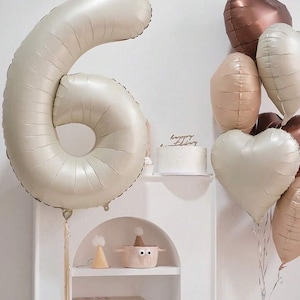 May include: A large, cream-colored, number six balloon with a string attached. The balloon is in front of a white shelf with a small cake on it. There are several heart-shaped balloons in the background, including a copper-colored heart balloon, a beige heart balloon, and a cream-colored heart balloon.