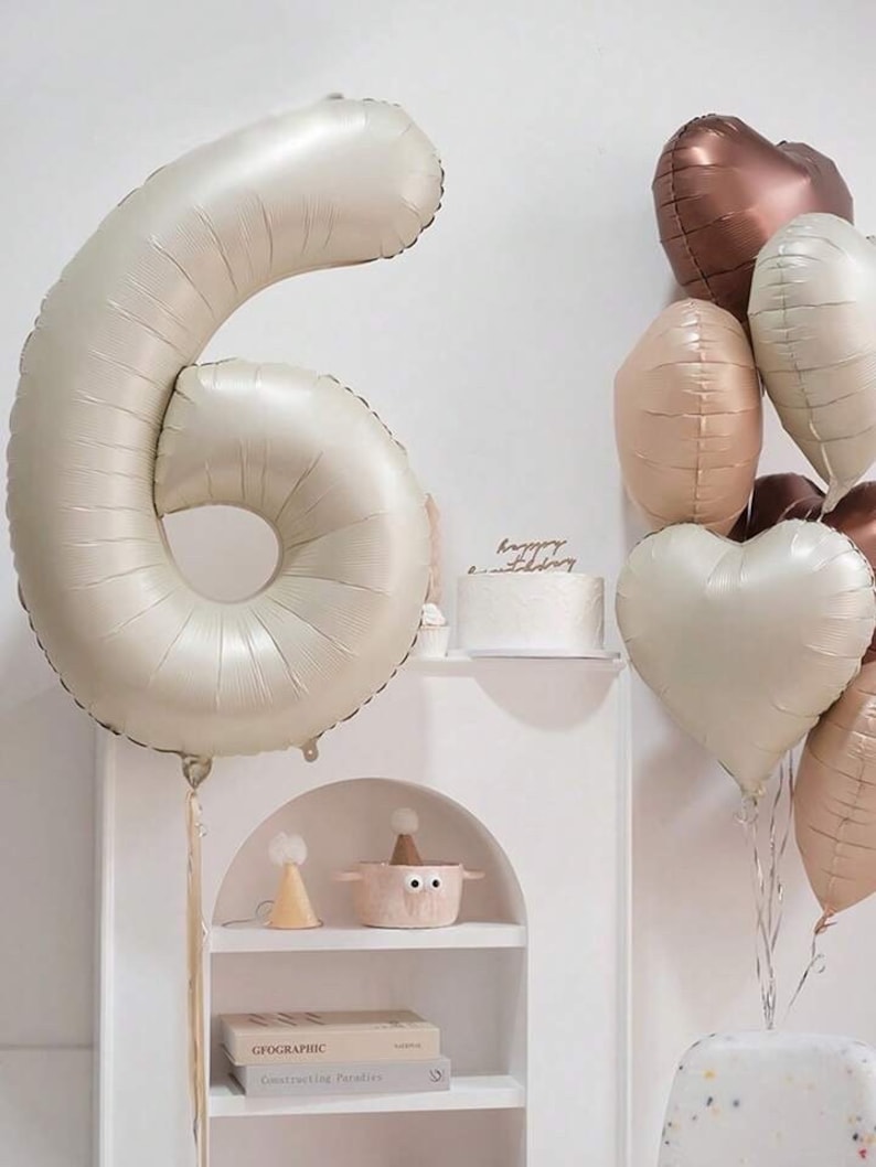 May include: A large, cream-colored, number six balloon with a string attached. The balloon is in front of a white shelf with a small cake on it. There are several heart-shaped balloons in the background, including a copper-colored heart balloon, a beige heart balloon, and a cream-colored heart balloon.