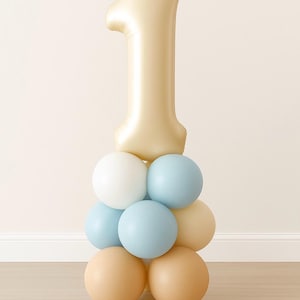 May include: A festive balloon arrangement for a first birthday celebration. A large gold number "1" balloon sits atop a pyramid of smaller balloons in white, light blue, and tan colors. The balloons are arranged on a light wood floor.