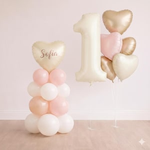 Puede incluir: Un conjunto de globos para una celebración de primer cumpleaños. Incluye un globo grande con el número uno, un globo en forma de corazón con el nombre "Sofia" y un ramo de globos en forma de corazón dorados, rosas y crema.