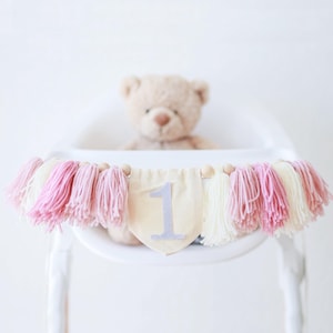 Puede incluir: Una guirnalda decorativa con borlas rosas, blancas y beige, y cuentas de madera. La guirnalda tiene un bander&iacute;n beige con el n&uacute;mero 1 en gris. Un osito de peluche est&aacute; sentado en una trona blanca.