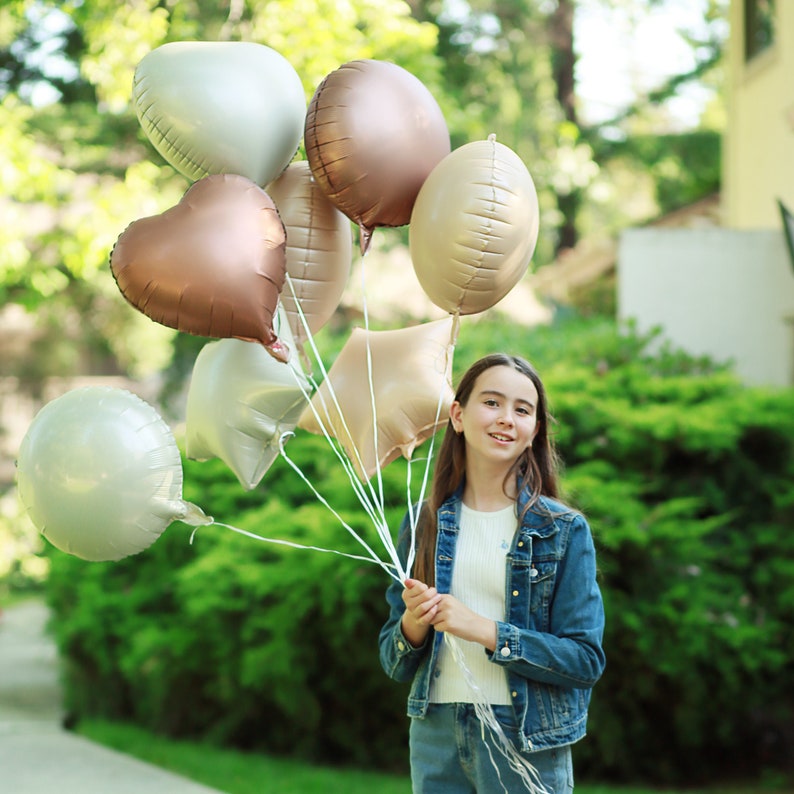 May include: A young person holding a bouquet of balloons in various shades of silver and brown. The balloons are in the shape of hearts and circles.