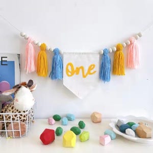 K&ouml;nnte beinhalten: Dekorative Girlande mit dem Wort &bdquo;One&ldquo; in gelber Schrift, umgeben von rosa, gelben und blauen Quasten. Die Girlande ist mit Holzperlen aufgereiht, passend f&uuml;r eine erste Geburtstagsfeier. Bunte Holzbl&ouml;cke sind darunter verstreut.
