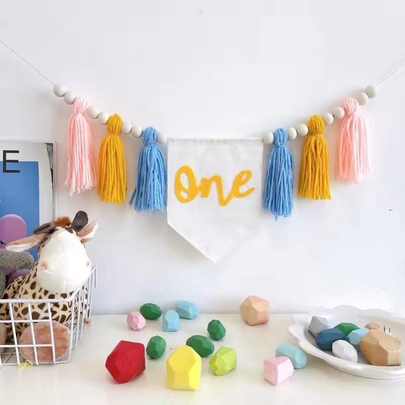 K&ouml;nnte beinhalten: Dekorative Girlande mit dem Wort &bdquo;One&ldquo; in gelber Schrift, umgeben von rosa, gelben und blauen Quasten. Die Girlande ist mit Holzperlen aufgereiht, passend f&uuml;r eine erste Geburtstagsfeier. Bunte Holzbl&ouml;cke sind darunter verstreut.