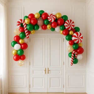 May include: A festive balloon archway in red, green, gold, and white, adorned with candy cane decorations. The balloons are arranged above a white double door, creating a celebratory entrance for a holiday event.