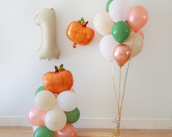 Può includere: Decorazioni per una festa di primo compleanno. Include un grande palloncino a forma di numero uno, palloncini a forma di zucca arancione e un bouquet di palloncini bianchi, verdi e pesca. L'insieme crea un'atmosfera festosa.