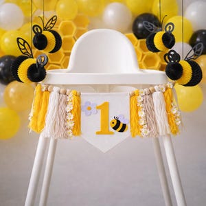 May include: A white high chair decorated for a first birthday. The banner reads "1" with a bee graphic. Yellow and black paper honeycomb bees hang from above. Yellow, white, and beige tassels and daisy accents adorn the banner.