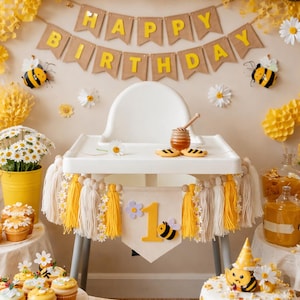 K&ouml;nnte beinhalten: Eine festliche Geburtstagsfeier mit gelben, wei&szlig;en und schwarzen Luftballons, G&auml;nsebl&uuml;mchen und Bienen-Dekorationen. Ein Hochstuhl ist mit Quasten und einem Banner mit der Aufschrift "1" geschm&uuml;ckt. Ein Kuchen und Cupcakes werden ausgestellt.