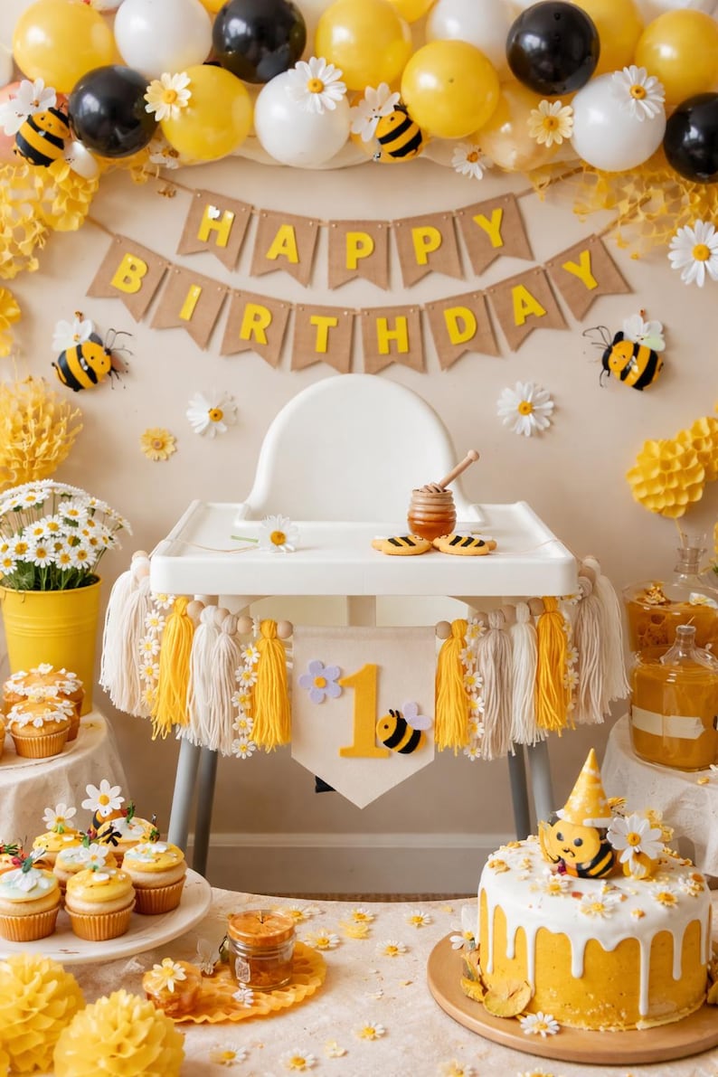 K&ouml;nnte beinhalten: Eine festliche Geburtstagsfeier mit gelben, wei&szlig;en und schwarzen Luftballons, G&auml;nsebl&uuml;mchen und Bienen-Dekorationen. Ein Hochstuhl ist mit Quasten und einem Banner mit der Aufschrift "1" geschm&uuml;ckt. Ein Kuchen und Cupcakes werden ausgestellt.