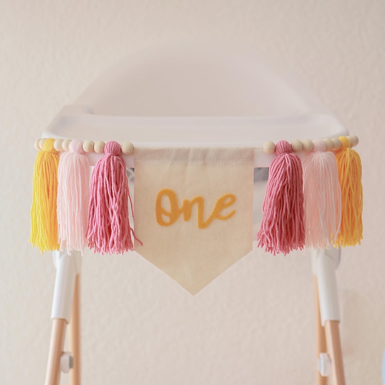 K&ouml;nnte beinhalten: Ein Hochstuhl-Banner mit dem Wort "One" in goldener Schrift auf einem cremefarbenen Wimpel. Das Banner ist mit rosa, gelben und pfirsichfarbenen Quasten und Holzperlen verziert. Das Banner ist an einem wei&szlig;en Hochstuhl befestigt.