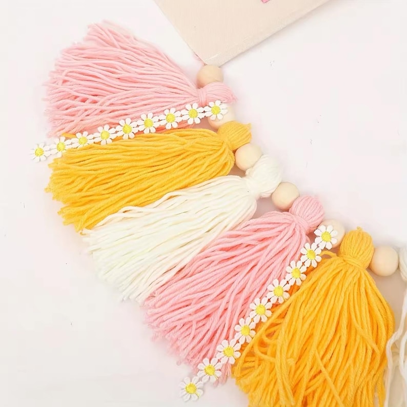 K&ouml;nnte beinhalten: Eine farbenfrohe Quasten-Girlande mit rosa, gelben und wei&szlig;en Garnquasten. Jede Quaste ist mit einer Kette aus kleinen G&auml;nsebl&uuml;mchen-Verzierungen und Holzperlen verziert. Diese dekorative Girlande ist ideal, um jedem Raum einen Hauch von Laune zu verleihen.