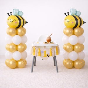 Peut inclure: Décoration de fête pour un premier anniversaire sur le thème des abeilles. Deux colonnes de ballons avec des ballons jaunes, dorés et blancs sont surmontées de ballons en forme d'abeille. Une chaise haute est décorée d'une bannière avec le chiffre "1" et une abeille.