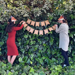 Op de afbeelding: Twee mensen met vossenmaskers hangen een banner op met de tekst "HAPPY BIRTHAY" tegen een achtergrond van groene klimop. Eén persoon draagt een rode jurk, de ander een groen overhemd en een zwarte broek. De banner is gemaakt van bruin papier.