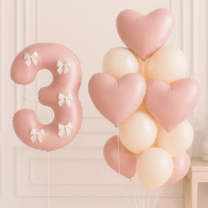 May include: A cluster of balloons in soft pink and cream colors. A large number "3" balloon is adorned with white bows. Heart-shaped and round balloons are tied together, creating a festive and celebratory atmosphere, perfect for a birthday or special event.