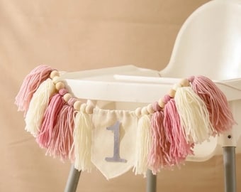 May include: A pink and white tassel garland with wooden beads and a white banner with the number "1" on it. The garland is hanging on a white high chair.