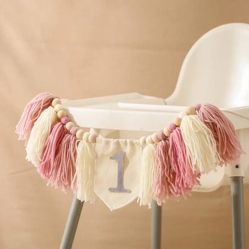 May include: A decorative banner for a high chair, featuring pink, cream, and wooden beads. The banner has a fabric pennant with a silver number 1, and is adorned with yarn tassels. Perfect for a first birthday celebration.