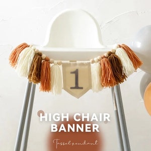 Puede incluir: Una guirnalda para trona con un colgante de borlas. La guirnalda presenta un bander&iacute;n de color neutro con el n&uacute;mero "1" y est&aacute; adornada con borlas crema, marr&oacute;n y naranja. Cuentas de madera ensartadas en la parte superior.
