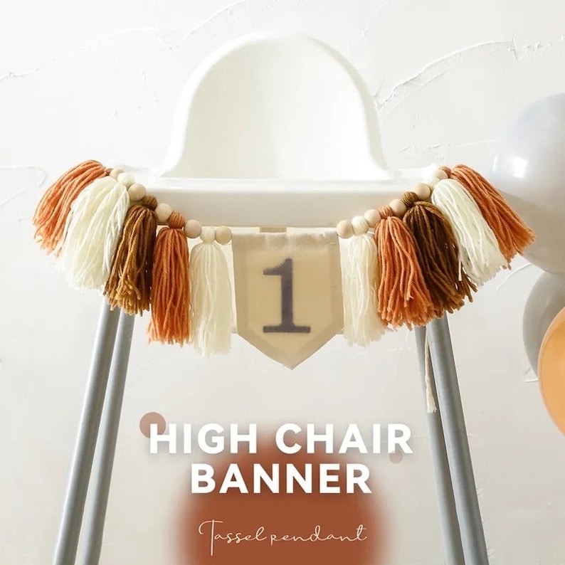Puede incluir: Una guirnalda para trona con un colgante de borlas. La guirnalda presenta un bander&iacute;n de color neutro con el n&uacute;mero "1" y est&aacute; adornada con borlas crema, marr&oacute;n y naranja. Cuentas de madera ensartadas en la parte superior.