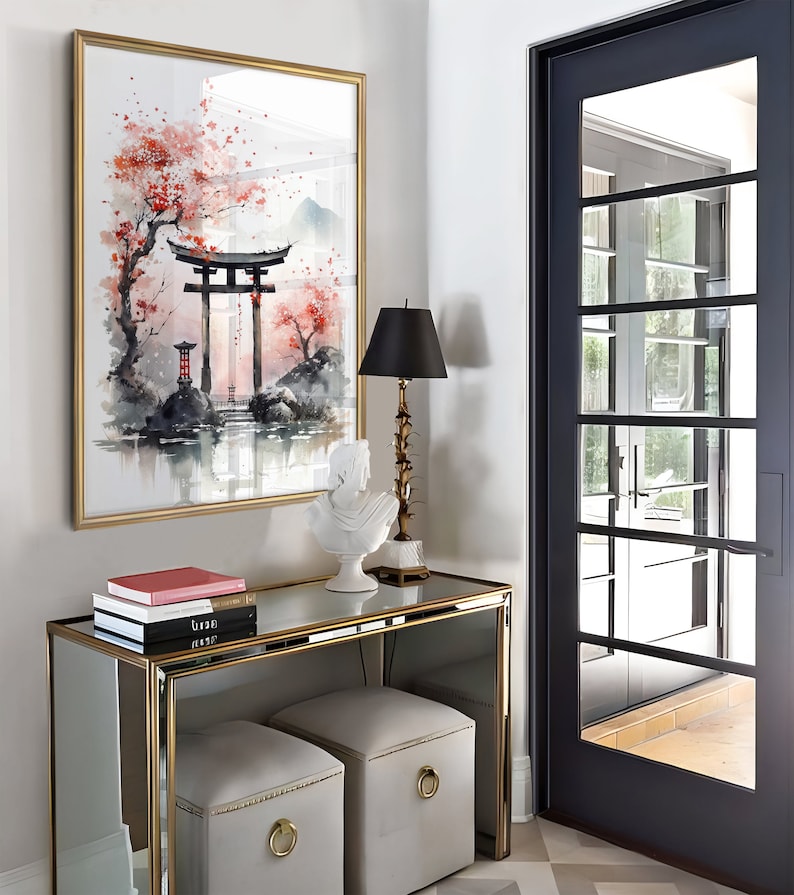 May include: A gold framed mirror with a watercolor painting of a Japanese garden scene. The painting features a red torii gate, a cherry blossom tree, and a small pond. A white ceramic sculpture of a bird sits on a gold mirrored console table with two gray upholstered ottomans. The console table has three books stacked on top of it.
