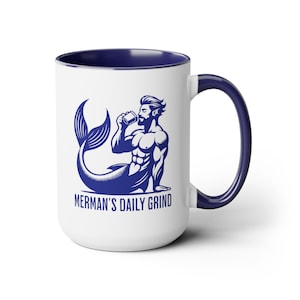 May include: White ceramic coffee mug with a blue rim and handle. The mug features a blue illustration of a merman drinking from a mug with the text "Merman's Daily Grind".