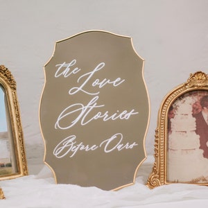 May include: A decorative sign with the text "the Love Stories Before Ours" in white script. The sign is a muted gold color with a gold border. Ornate gold picture frames with photos are in the background.