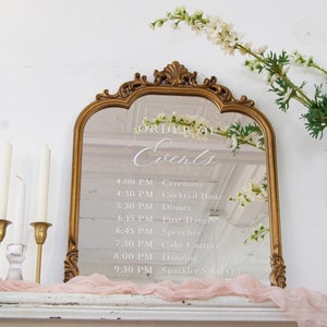 May include: A gold-framed mirror with an ornate design, displaying a white vinyl sticker with the text "ORDER OF Events" and a list of wedding events with times. The mirror is sitting on a white surface with a pink fabric draped over it.