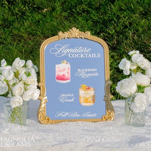 May include: A gold-framed sign with the words "Signature Cocktails" and illustrations of two cocktails: a Blackberry Margarita and an Orange Crush. The sign is on a white tablecloth with floral arrangements on either side.
