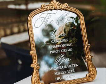 May include: A gold-framed mirror with ornate detailing displays a bar menu. The text includes "The Bar," followed by wine and beer selections: Cabernet, Chardonnay, Pinot Grigio, Michelob Ultra, Miller Lite, and High Noon. The mirror is set on a white surface.