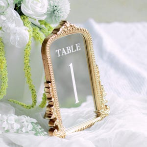 May include: Gold table number holder with a mirrored surface. The holder has an ornate frame and the number "1" is displayed in the mirror. The text "TABLE" is printed above the number.