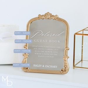 A gold wedding mirror being used as decoration for a wedding entrance table that has a polaroid guest book on it, with the wedding mirror having custom vinyl decal words on it that prompt wedding guests to leave a polaroid and a message as an alternative wedding guest book ideas.