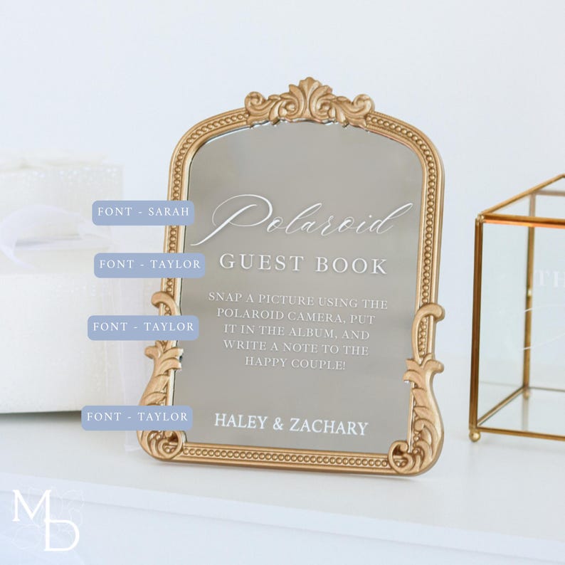 A gold wedding mirror being used as decoration for a wedding entrance table that has a polaroid guest book on it, with the wedding mirror having custom vinyl decal words on it that prompt wedding guests to leave a polaroid and a message as an alternative wedding guest book ideas.