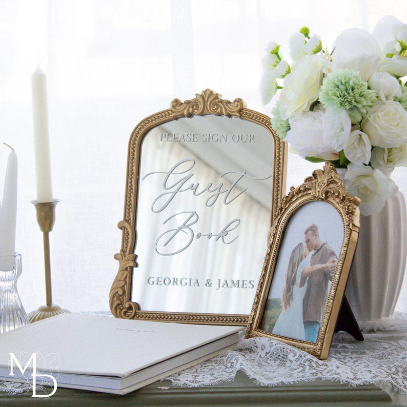 A gold wedding mirror being used as decoration for a wedding entrance table that has a polaroid guest book on it, with the wedding mirror having custom vinyl decal words on it that prompt wedding guests to leave a polaroid and a message as an alternative wedding guest book ideas.
