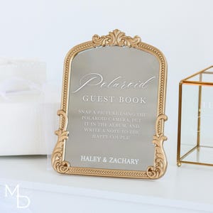 A gold wedding mirror being used as decoration for a wedding entrance table that has a polaroid guest book on it, with the wedding mirror having custom vinyl decal words on it that prompt wedding guests to leave a polaroid and a message as an alternative wedding guest book ideas.