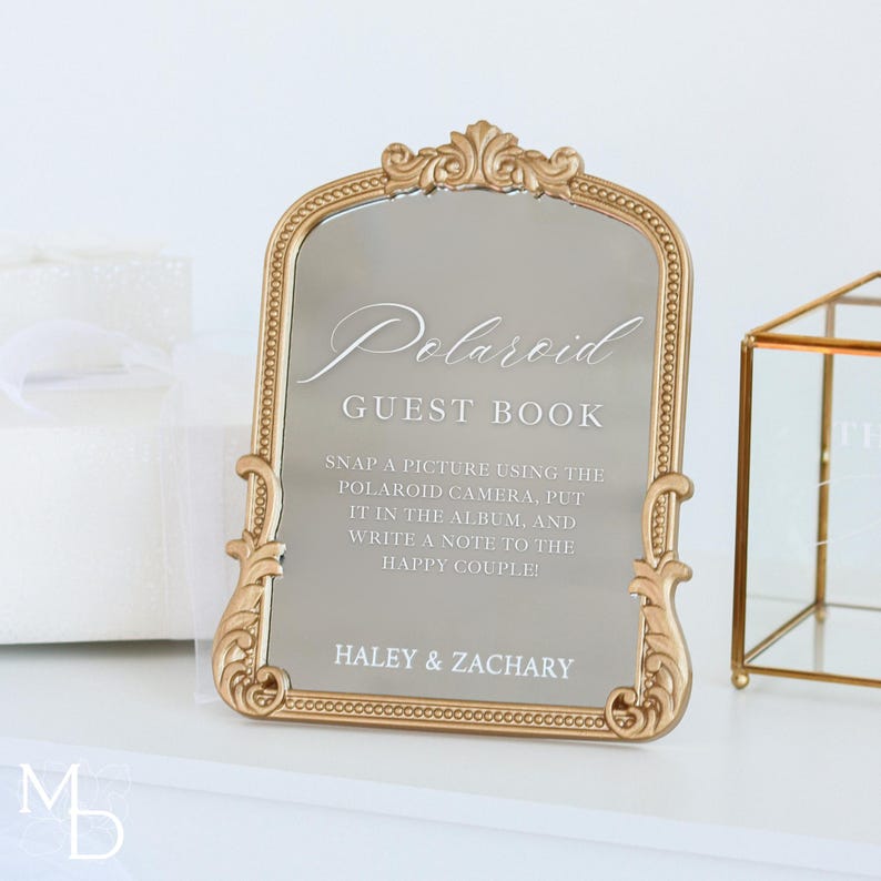 A gold wedding mirror being used as decoration for a wedding entrance table that has a polaroid guest book on it, with the wedding mirror having custom vinyl decal words on it that prompt wedding guests to leave a polaroid and a message as an alternative wedding guest book ideas.