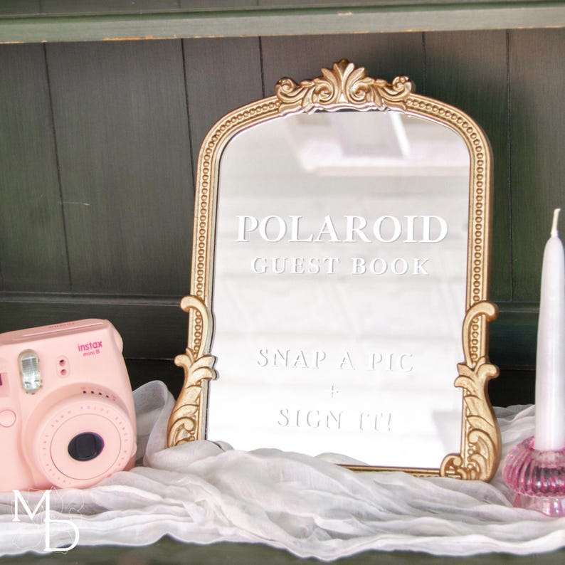 A gold wedding mirror being used as decoration for a wedding entrance table that has a polaroid guest book on it, with the wedding mirror having custom vinyl decal words on it that prompt wedding guests to leave a polaroid and a message as an alternative wedding guest book ideas.
