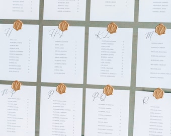 May include: A set of white cards with gold wax seals and handwritten names on each card. The cards are arranged in rows on a mirrored surface.