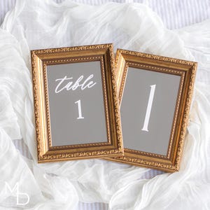 May include: Two gold framed mirrors with a gray background. The left mirror has the text "table 1" written in white. The right mirror has the number "1" written in white.