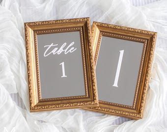 May include: Two gold framed mirrors with a gray background. The left mirror has the text "table 1" written in white. The right mirror has the number "1" written in white.