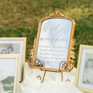 May include: A gold-framed mirror with the text "Wish you were here" and a message of remembrance. The mirror is displayed on a stand, surrounded by framed photos, likely for a memorial or wedding.